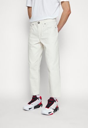 Osoba nosząca białe skrócone jeansy, białą koszulkę i białe, czarne oraz czerwone buty sportowe, stojąca na jednolitym białym tle.