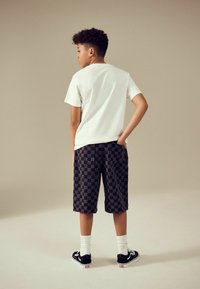Hvid t-shirt med korte ærmer, shorts i sort og grå tern, hvide ankelstrømper, sorte sneakers med hvide detaljer. Barnet vender væk.