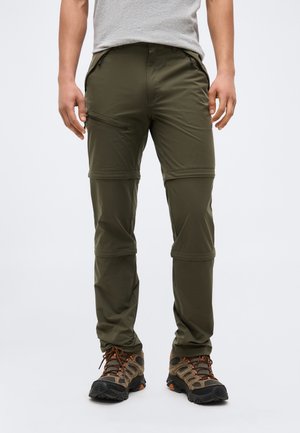 Egy férfi olívazöld, átalakítható túranadrágot és barna túracipőt visel, egyszerű háttér előtt áll.