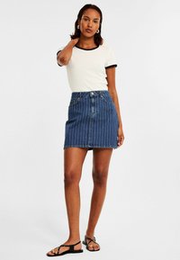 Weißes Hemd mit schwarzem Rand, kombiniert mit einem blauen Jeansrock mit vertikalen weißen Streifen. Das Model steht mit sichtbaren schwarzen Sandalen.