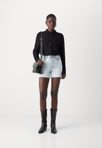 Camicia nera a bottoni, pantaloni di jeans corti con bordi sfrangiati e stivali neri con tacco. Gli accessori comprendono una piccola borsa a tracolla nera.