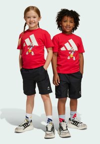 Červené bavlněné trička mají velké bílé logo Adidas a kreslenou postavičku, v kombinaci s černými šortkami. Oba mají na sobě tenisky s výraznými designy.