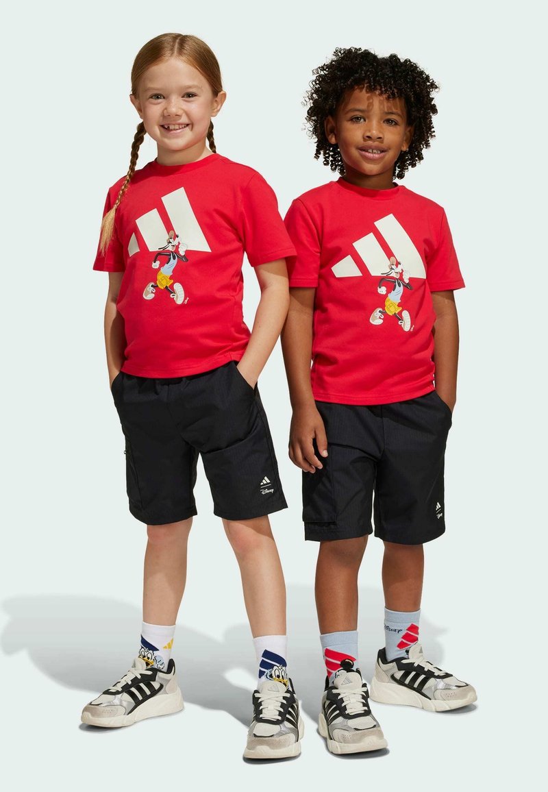 Červené bavlněné trička mají velké bílé logo Adidas a kreslenou postavičku, v kombinaci s černými šortkami. Oba mají na sobě tenisky s výraznými designy.