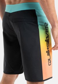 Bañador negro con una cinturilla azul clara, con un panel lateral de degradado de naranja a verde y un bolsillo trasero; logo impreso en un lado.