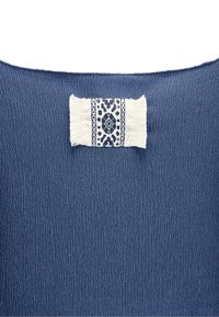 Niebieska, teksturowana tkanina z wovewą metką z wzorem w białym i granatowym, otoczoną postrzępionymi brzegami. Metka umieszczona centralnie na górze.