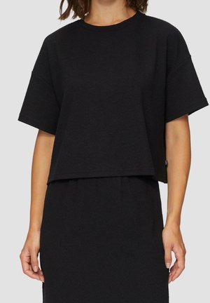 Frau trägt ein schlichtes schwarzes, kurzärmeliges, bauchfreies Oberteil und einen passenden hoch taillierten Rock vor einem hellgrauen Hintergrund.