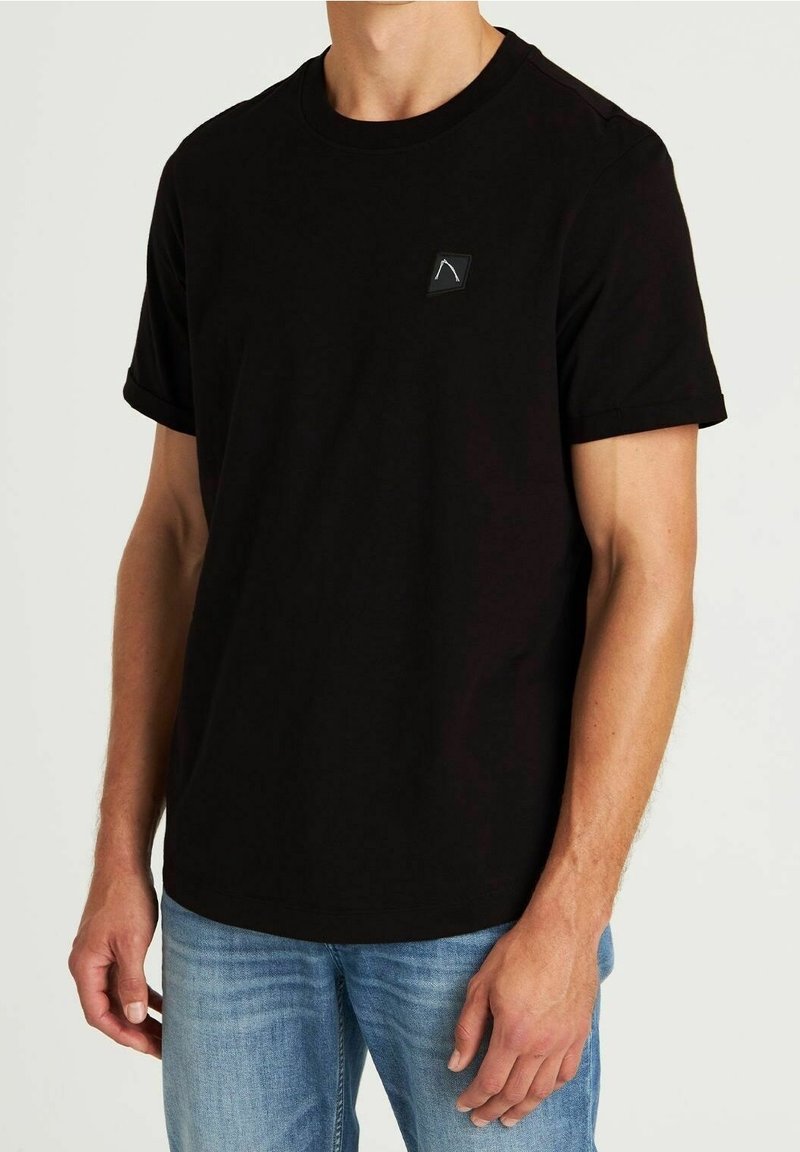Homme portant un t-shirt noir uni à manches courtes avec un petit logo blanc sur la poitrine et un jean bleu clair, debout devant un fond blanc.