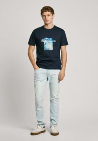 Camiseta de algodón de color marino con un estampado gráfico azul y el texto "Pepe Jeans". Combinada con unos jeans de color azul claro y unas zapatillas blancas con suelas marrones.
