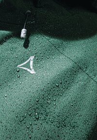 Grüne wasserdichte Jacke mit strukturierter Oberfläche, die sichtbare Wassertropfen aufweist und ein weißes Logo auf der Brustpartie hat.