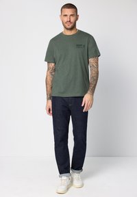 Olivově zelené bavlněné tričko s krátkými rukávy a potiskem loga na prsou, kombinované s tmavě modrými džínami a bílými teniskami.