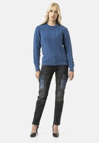 Pull en tricot bleu avec un motif de câble en losange ; associé à un pantalon cargo noir avec des accents bleus et plusieurs poches ; talons hauts.