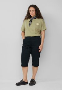 Olijfgroene T-shirt met korte mouwen en een zonafbeelding, gecombineerd met donkere, cropped broek en zwarte instappers met zilverkleurige hardware-details.