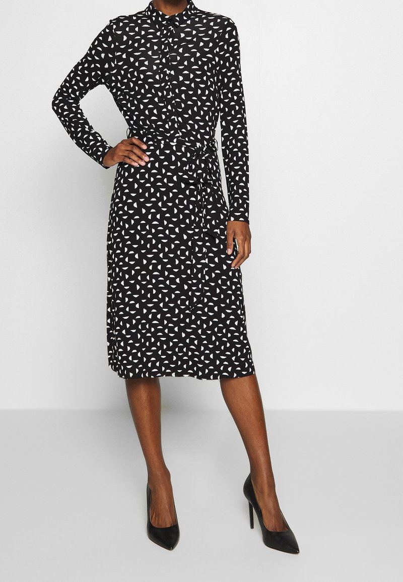 Svart klänning med ett geometriskt vitt mönster, långa ärmar, krage och ett knytband i midjan. Bärs med svarta högklackade skor.