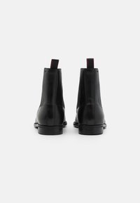 Bottes noires en cuir à cheville avec une texture lisse, des panneaux latéraux élastiques et de petites languettes rouges à l'arrière. Semelles plates pour la stabilité.