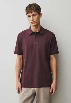 Noor mees hele naha ja lühikeste pruuni juustega, kellel on seljas tumelilla lühikeste varrukatega polo-särk ja heledad beežid püksid, seisab tavalise seina ees.