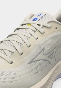 Κοντινό πλάνο ενός μπεζ αθλητικού παπουτσιού με πάνελ από δίχτυ, λευκά κορδόνια και μπλε λογότυπο στη γλώσσα και λεπτομέρειες στην πλευρά.