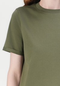 Osoba nosząca gładką oliwkowozieloną koszulkę z krótkim rękawem i podwiniętymi mankietami, pokazana od szyi do środkowej części tułowia na jasnym tle.