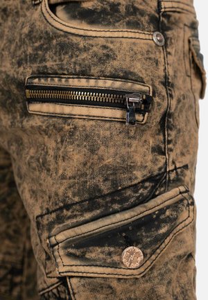 Cargo pants in distressed beige denim, featuring a textured dark pattern, zippered pockets, and metal hardware accents.