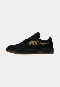 Zapatilla baja de color negro y marrón con cierre de cordones, cuello acolchado y suela con marca sobre un fondo blanco.