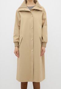 Abrigo largo beige con cierre de doble botonadura, gran capucha y sutiles detalles en los botones. Presenta puños elásticos y bolsillos laterales. Textura suave.