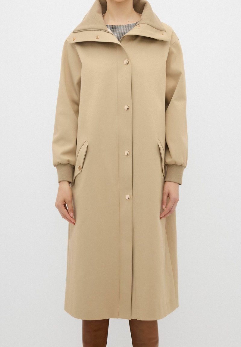 Abrigo largo beige con cierre de doble botonadura, gran capucha y sutiles detalles en los botones. Presenta puños elásticos y bolsillos laterales. Textura suave.
