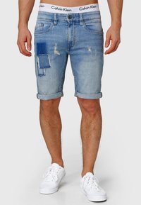 Shorts en denim bleu clair présentant un design déchiré avec des ourlets retournés et une accent de patch. Portés avec des baskets blanches.