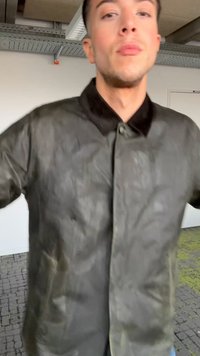 Jonge man die een donker, licht glanzend jack met een zwarte kraag draagt, staand binnen in een kamer met industriële plafondbuizen.