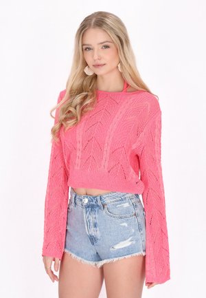 Junge Frau mit langen blonden Haaren, die einen rosa Strickpullover und hellblaue Jeansshorts trägt, steht vor weißem Hintergrund.