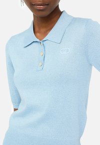 Polo en tricot bleu clair à manches courtes, avec un col, trois boutons beige et un logo brodé sur la poitrine.