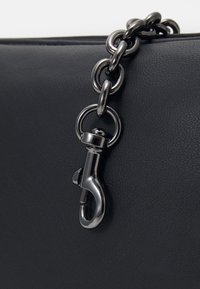 Detail černé kožené tašky s kovovým řetízkem a zapínáním, se strukturovaným povrchem a lesklým kováním.