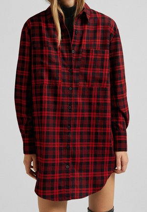 Kobieta w długiej czerwono-czarnej koszuli w kratę na guziki z dwoma kieszeniami na piersi i długimi rękawami, z rękami swobodnie opuszczonymi wzdłuż ciała.
