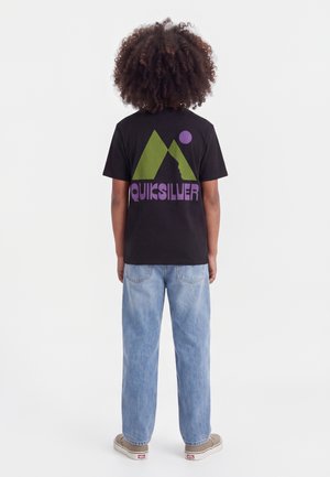 Enfant aux cheveux bouclés portant un t-shirt noir Quiksilver avec un logo vert et violet, un jean bleu clair et des baskets beiges, debout, de dos.