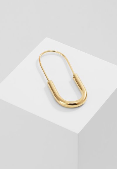 Kultainen metallinen turvaklipsi, jossa on sileä, pyöreä muoto ja kiillotettu pinta, lepää valkoisella geometristä alustalla.