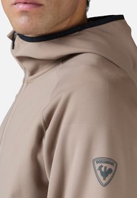 Beige hættejakke med en glat tekstur og syning detaljer. Viser et blåt Rossignol-logo på højre arm.