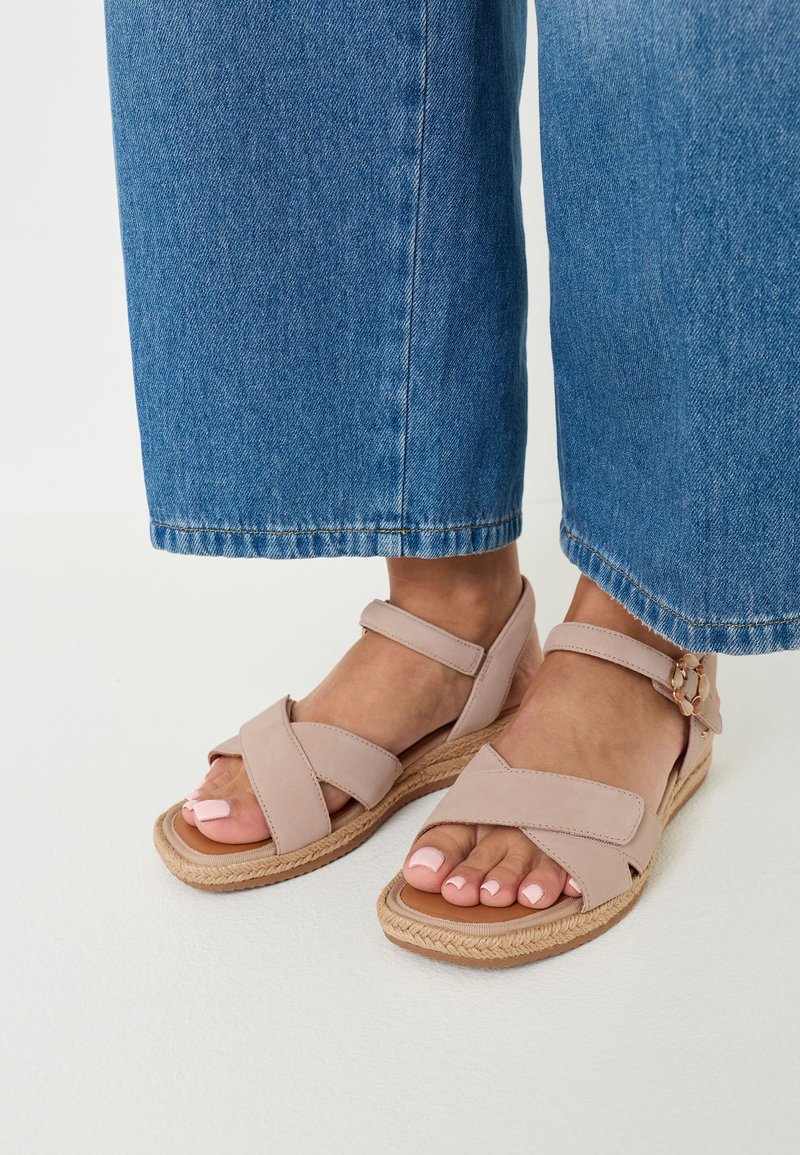 Pieds en sandales roses claires à brides croisées avec boucle à la cheville et semelle style espadrille, portant un pantalon en denim bleu à jambes larges coupé au-dessus des chevilles.
