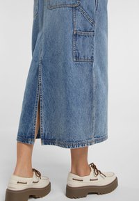 Falda larga de mezclilla con una abertura lateral, características de bolsillos, dobladillo deshilachado y lavado azul. Combinada con zapatos beige y cordones marrones, sobre una suela texturizada.