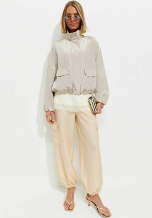 Femme portant une veste bomber beige clair, pantalon ample crème, sandales dorées à talons hauts, lunettes de soleil, tenant une petite pochette.
