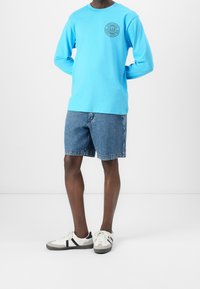 Hombre vestido con una camisa de manga larga azul claro con logo, pantalones cortos de mezclilla y zapatillas blancas, de pie contra un fondo blanco.