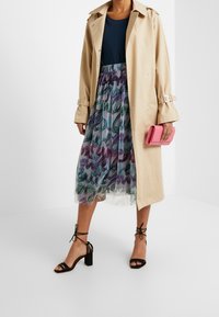 Trench coat beige, blusa azul, falda translúcida con patrones abstractos en verde y púrpura, sandalias de tacón negro y clutch rosa.