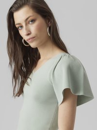 Mintgrønn topp med ribbestruktur, V-hals og puffede korte ermer. Modellen har langt hår og sølvfargede hoopøreringer. Nøytral bakgrunn.