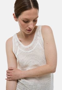 Haut en crochet sans manches blanc avec un col rond. Présente un motif de maille ouverte texturé et une silhouette ajustée.
