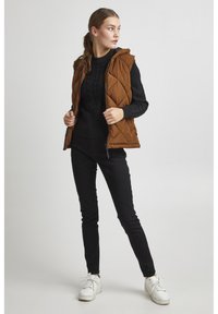 Gilet rembourré marron avec capuche, porté sur un pull en maille torsadée noir. Un pantalon noir et des baskets blanches complètent la tenue.