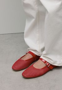Ballerines en maille rouge avec une bride rouge brillante et un détail de boucle. Associées à un pantalon blanc, mettant en avant un design simple, plat et arrondi.