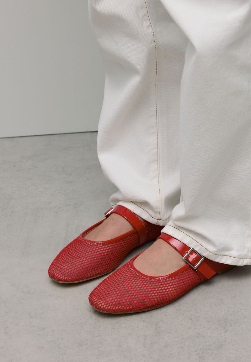 Ballerines en maille rouge avec une bride rouge brillante et un détail de boucle. Associées à un pantalon blanc, mettant en avant un design simple, plat et arrondi.