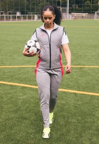 Moteris pilku ir raudonu sportiniu kostiumu, laikanti futbolo kamuolį, eidama dirbtinės žolės aikšte futbolo stadione, fone matyti vartai.