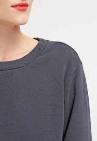 Felpa grigia scura con colletto a coste, texture morbida e vestibilità rilassata; presenta un sottile dettaglio di cucitura sulle spalle.