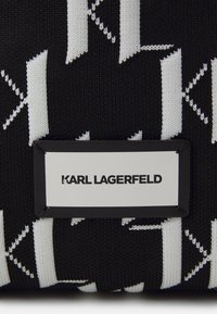 Czarna torba z teksturowanej tkaniny z białym wzorem oraz prostokątną białą metką z napisem "KARL LAGERFELD" w pogrubionej czarnej czcionce.