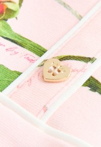 Tissu rose avec un motif floral agrémenté d'accents verts, détaillé avec une étiquette en forme de cœur en métal doré. La texture semble lisse.