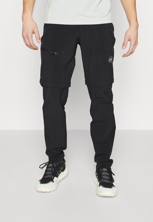 Mann trägt schwarze Zip-off-Hose und schwarz-weiße Sneaker, steht vor einfachem weißem Hintergrund.