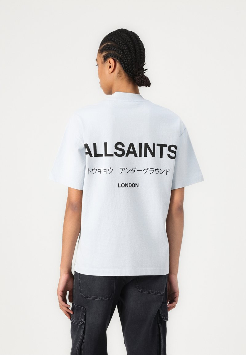 Lyseblå bomull T-skjorte med løs passform, med en stor svart "ALLSAINTS" logo og japansk tekst på baksiden. Sorte bukser er brukt.
